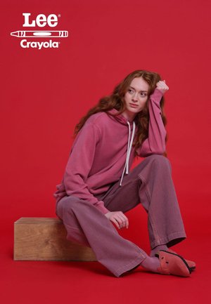Pink hoodie with drawstrings, striped wide-leg trousers, and pink slippers. Model seated on a wooden block against a solid red background.