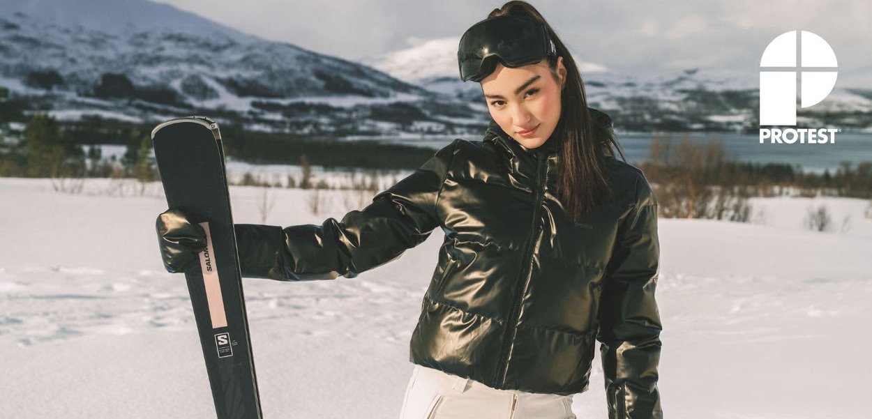 Glanzend zwarte donsachtige ski-jas, witte sneeuwbroek, een zwarte ski met roze accenten vasthoudend, zwarte skibril dragend, besneeuwde bergachtergrond.
