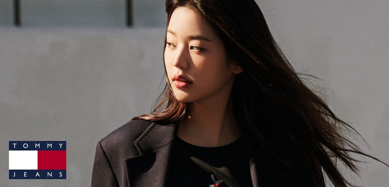 Modelo vistiendo un blazer negro sobre una camiseta gráfica. El cabello es largo y fluido. El fondo es neutro con una iluminación suave. La marca es visible en la esquina.