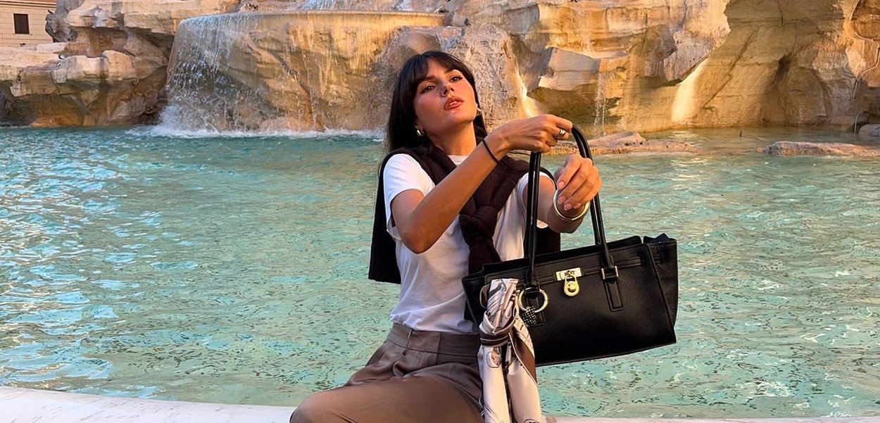 Giovane donna con t-shirt bianca e pantaloni marroni, che tiene una borsa nera, seduta vicino a una fontana di pietra con acqua che scorre.
