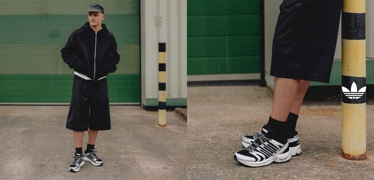 Giacca nera con zip, camicia bianca, pantaloncini neri e calzini neri con scarpe da ginnastica bianche con dettagli argentati e neri. Sfondo in cemento.