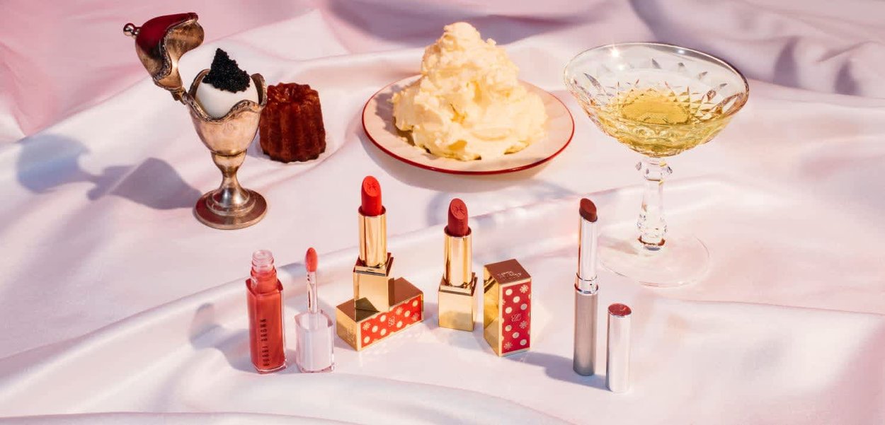 Une collection de produits de beauté comprenant des rouges à lèvres rouges avec un emballage doré, un rouge à lèvres liquide rose, un verre en cristal et des desserts sur un tissu en satin.