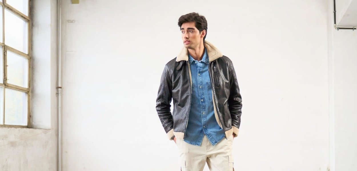 Giubbotto bomber in pelle nera con colletto in shearling, indossato su una camicia di denim chiaro e pantaloni cargo beige, su uno sfondo neutro.