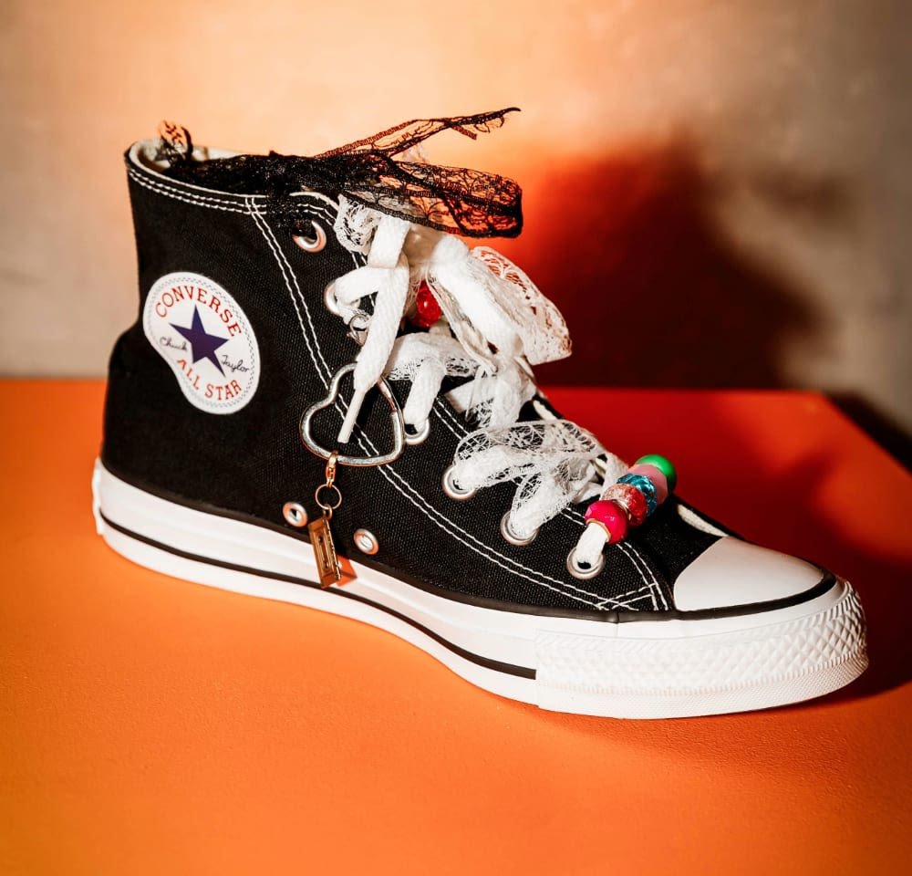 Black high-top sneaker with white sole, lace accents, decorative beads on laces, and Converse logo patch. Textured canvas material.
