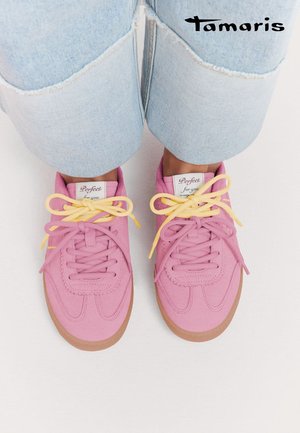 Rosa Turnschuhe mit gelben Schnürsenkeln, getragen zu hochgekrempelten hellblauen Jeans auf weißem Hintergrund, Markenlogo "Tamaris" sichtbar.