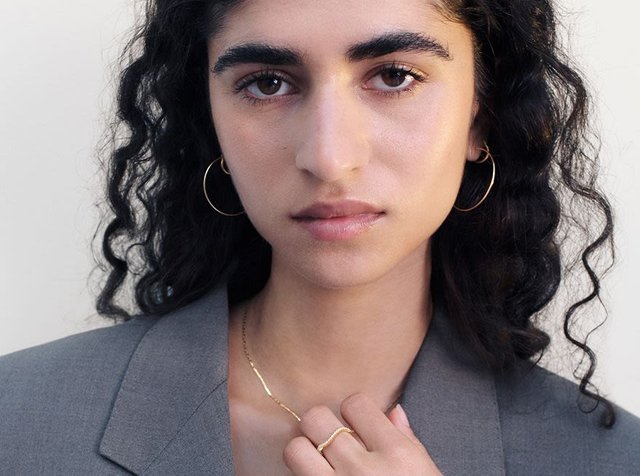 Guld hoop øreringe og en delikat guldring båret med en grå blazer. Emnet har krøllet sort hår og neutral makeup.