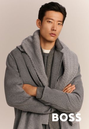 Szary wełniany płaszcz z fakturowanym, dzianinowym kołnierzem typu szal, nałożony na szary sweter z zamkiem błyskawicznym. Model stoi z założonymi rękami na neutralnym tle.