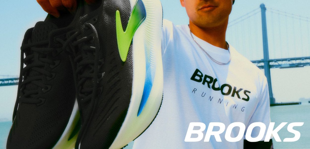 Scarpe da corsa nere con accenti verdi e suole blu, caratterizzate da una tomaia testurizzata e lacci, tenute da una persona con una maglietta bianca Brooks.