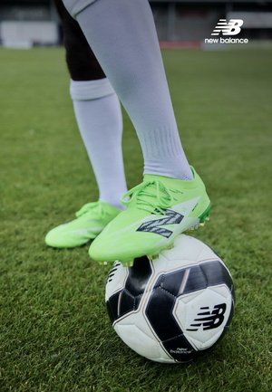 Scarpette da calcio verde brillante con tomaia testurizzata sono posizionate su un pallone da calcio bianco e nero sull'erba. Il logo di New Balance è visibile su entrambi.