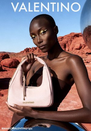 Modelka z makijażem oczu w kolorze złota trzyma bladoróżową torebkę Valentino na tle czerwonych skał pustyni i czystego błękitnego nieba.