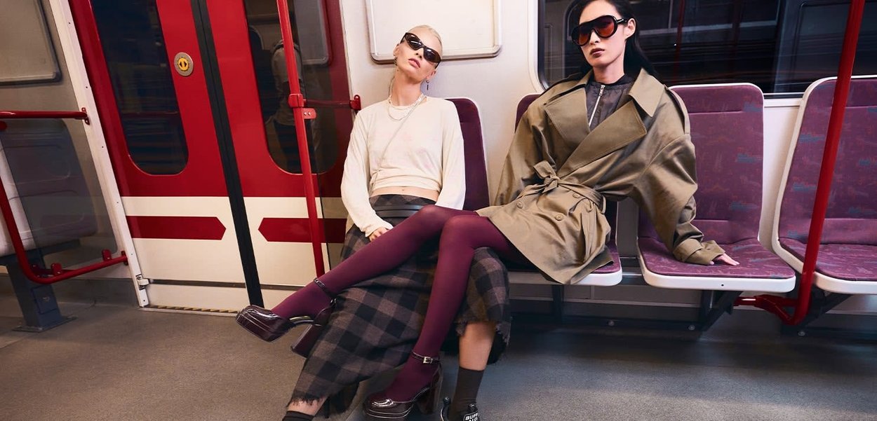 Two models sit on a subway bench. One wears a light-colored cropped top and plaid skirt; the other dons a long olive trench coat. Both have sunglasses.