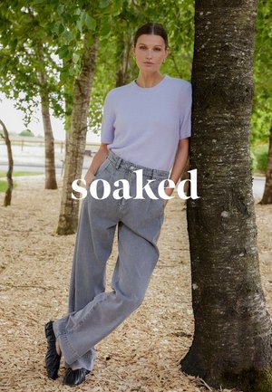 Donna con maglione lavanda a maniche corte e pantaloni grigi larghi appoggiata a un albero in un'area boschiva all'aperto con sovrapposizione di testo "soaked".