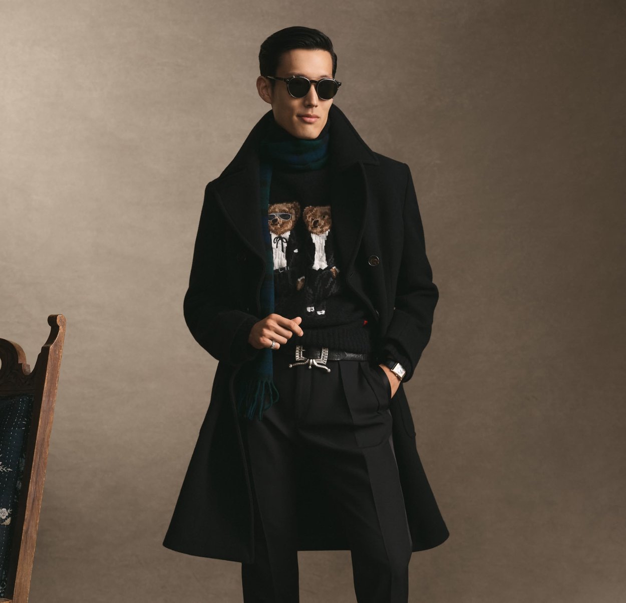 Svart dubbelknäppt rock över en mörk tröja med björnmotiv, kombinerad med skräddarsydda svarta byxor och en grön scarf. Har runda solglasögon.