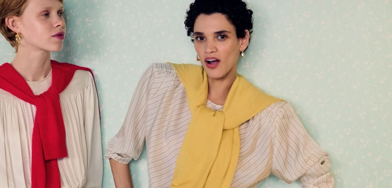 To kvinder står op ad en lyst mønstret væg iført stribede bluser med trøjer hængt over skuldrene, den ene rød og den anden gul.