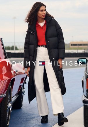 Abrigo acolchado negro, camiseta polo roja, pantalones de pierna ancha blancos y botas de tobillo negras. Coches visibles en el fondo. Entorno al aire libre.