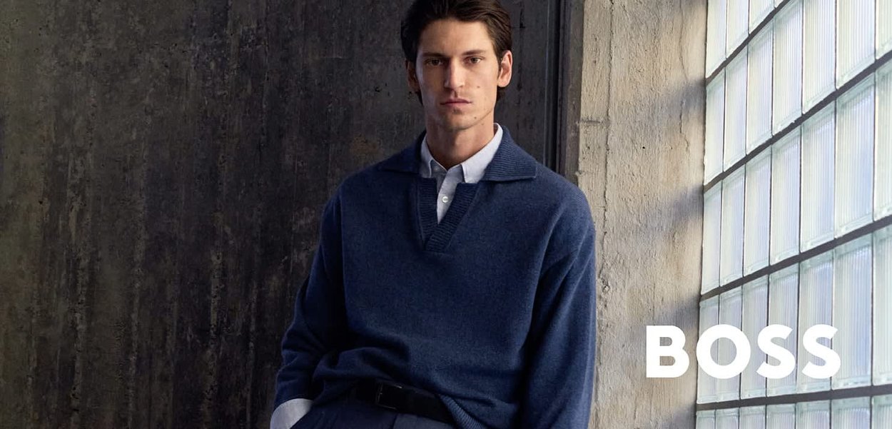 Uomo che indossa un maglione blu navy e una camicia bianca seduto contro una parete scura vicino a una finestra con la scritta "BOSS" su blocchi di vetro.