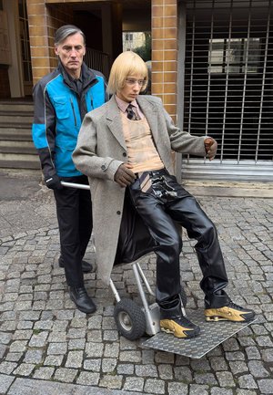 Ein Mann mit schwarzen Handschuhen schiebt einen Rollwagen, auf dem eine Schaufensterpuppe mit einem Hahnentrittmantel, Lederhose und Sonnenbrille steht, über Kopfsteinpflaster.