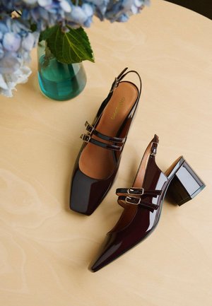 Talons slingback en cuir verni bordeaux avec un bout carré, deux sangles à boucle et un talon bloc épais, sur une surface en bois clair.