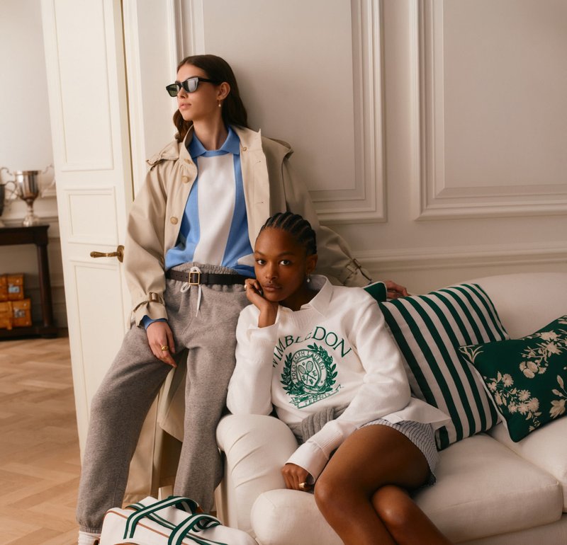 Dos mujeres con ropa informal posan en el interior, una de pie con gafas de sol y gabardina, la otra sentada en un sofá con cojines a rayas.