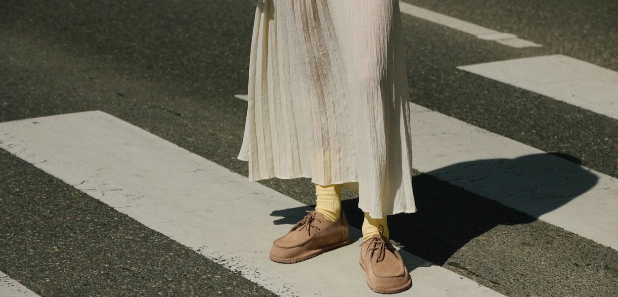 Beige suede shoes with lace detailing, worn with yellow ribbed socks, paired with a light pleated dress, standing on a crosswalk.