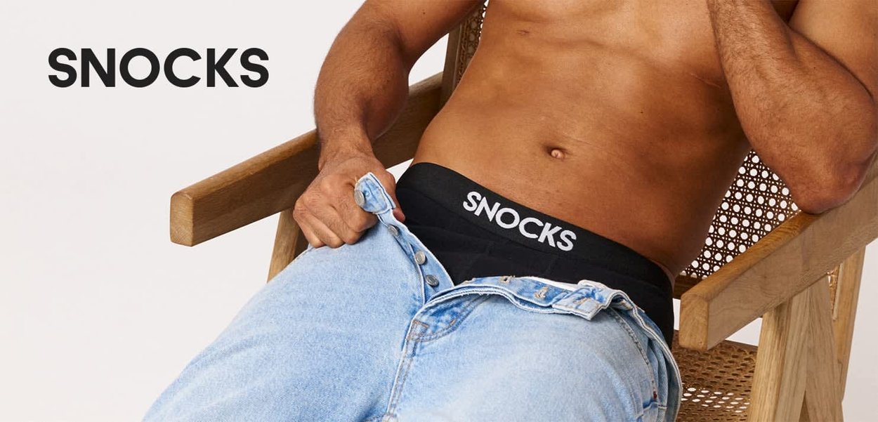 Skjorteløs person sidder i en stol og åbner lyseblå jeans, iført sort undertøj med Snocks-logo på taljebåndet.