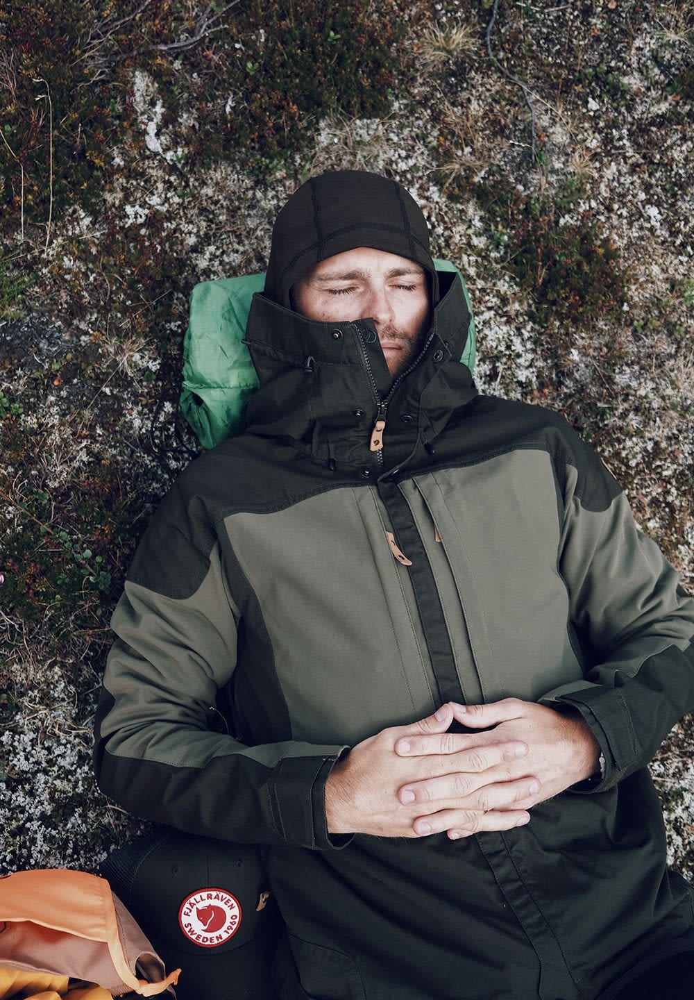 Grön och svart jacka med hög krage, dragkedjor och fickor; bärs av en person som ligger på en stenig yta. Brun logotyp på en cap i närheten.