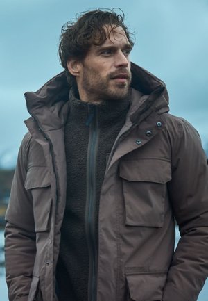 Brauner wasserdichter Anorak mit Kapuze, der mehrere Taschen und einen Reißverschluss aufweist. Darunter ist ein dunkler, strukturiertes Fleece-Oberteil sichtbar.