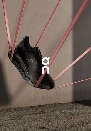 Scarpa da ginnastica nera sospesa tra fasce elastiche rosa, con una tomaia testurizzata e una suola ammortizzata con bordi arrotondati. Logo visibile.