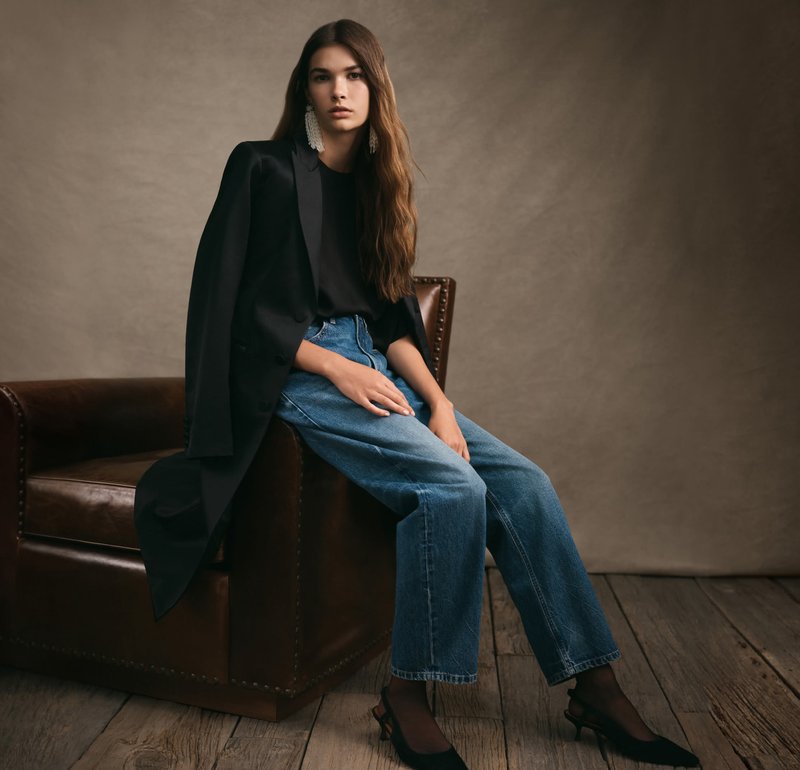 Blazer noir ajusté posée sur l'épaule, accompagné d'un haut noir, d'un jean bleu taille haute et de chaussures à talons noires, assise sur une chaise en cuir.