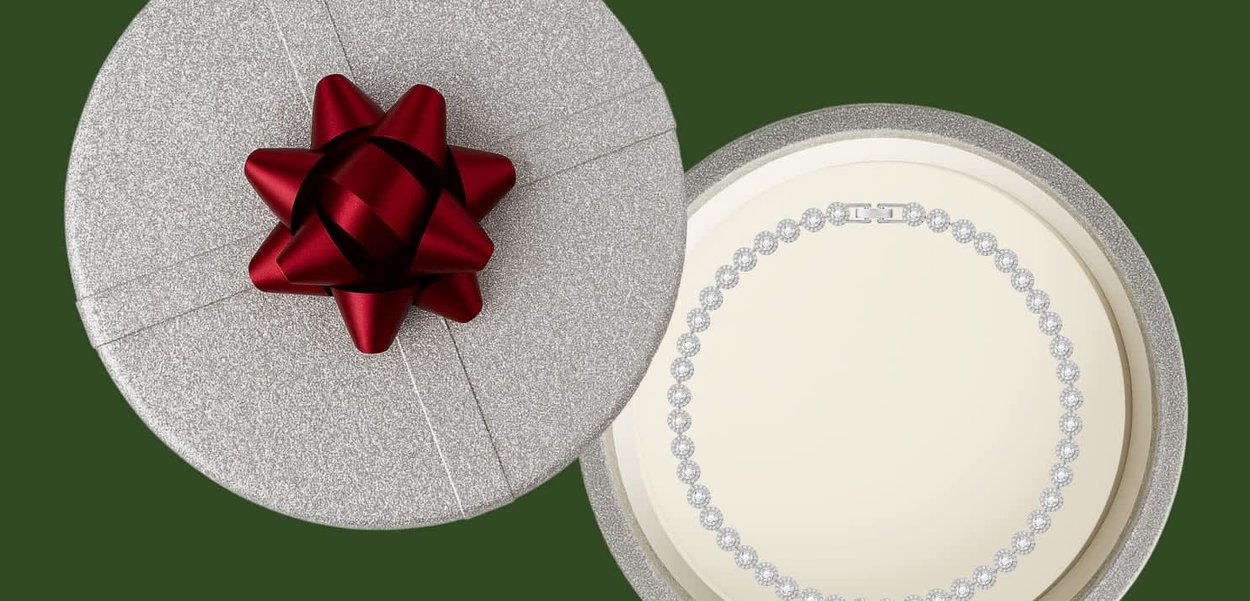 Ronde zilveren geschenkdoos met een rode strik, half open, waarin een diamanten tennisketting op een crèmekleurige achtergrond zichtbaar is.