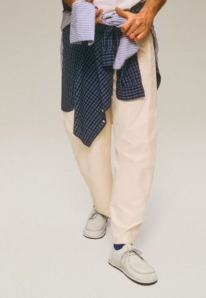 Person, die ein blau kariertes Hemd um cremefarbene Hosen bindet, weiße Schnürschuhe und blaue Socken trägt, vor einem schlichten Hintergrund steht.