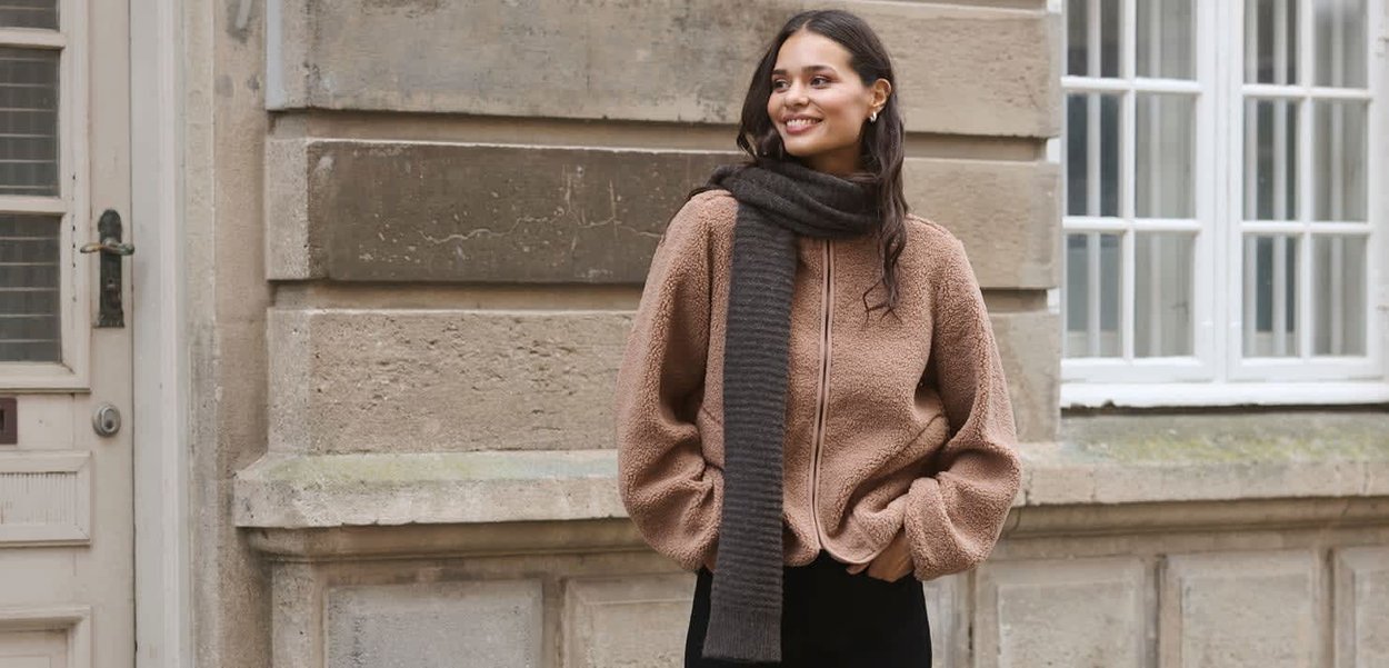 Ruskea fleece-zip-up-takki, jossa on ylisuuret hihat, yhdistettynä tummanharmaaseen neulehuiviin. Malli seisoo kiviseinää vasten.