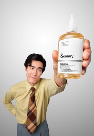 Mężczyzna w żółtej koszuli i wzorzystym krawacie wyciąga butelkę toniku złuszczającego The Ordinary Glycolic Acid 7% na jednolitym tle.