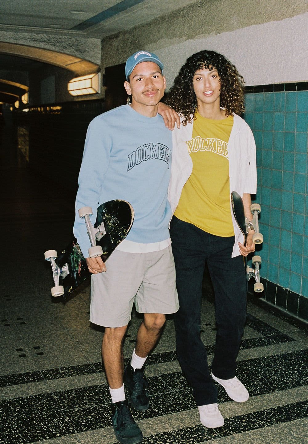 Deux personnes se tiennent dans un couloir, chacune tenant un skateboard. La personne à gauche porte un sweat-shirt bleu clair et un short beige. La personne à droite porte une chemise jaune avec une veste blanche et un pantalon sombre.