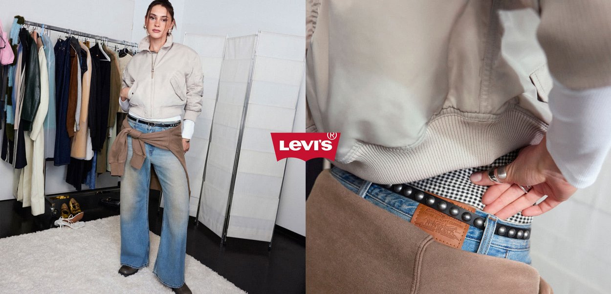 Woman wearing beige bomber jacket, blue wide-leg jeans, and brown sweater tied at waist, standing by clothes rack in a fitting room.