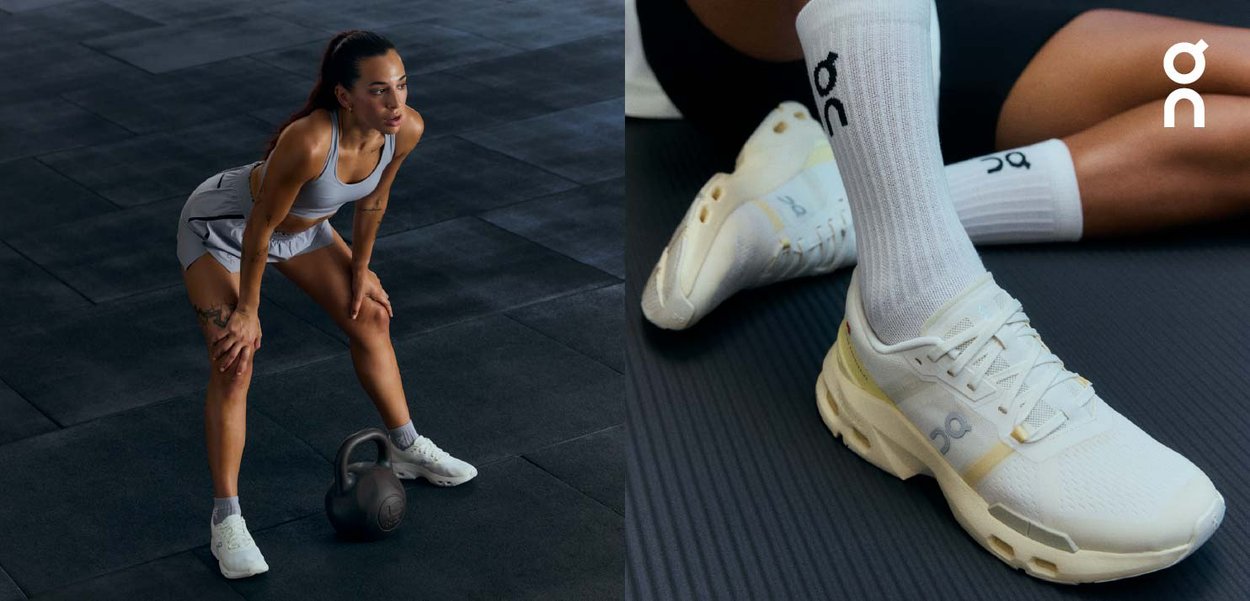 Kvinna i grå träningskläder vilar händerna på böjda knän bredvid en kettlebell, iförd vita On-skor och vita strumpor på svart golv.