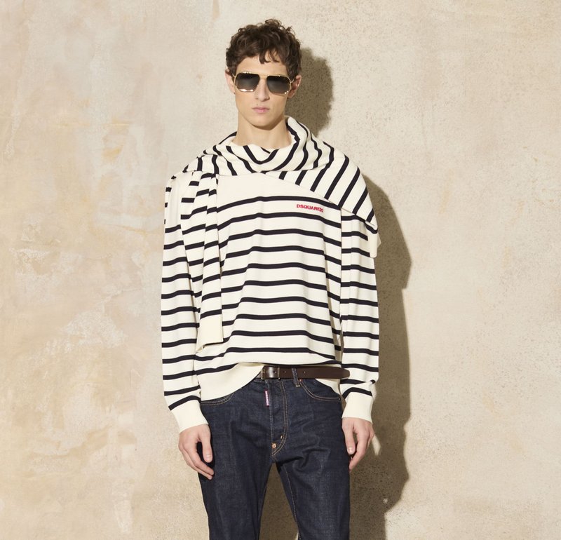 Gestreifter Langarm-Sweatshirt in Creme und Marineblau, mit lockerer Passform und rotem Logo; kombiniert mit dunklen Denim-Jeans und Sonnenbrille.