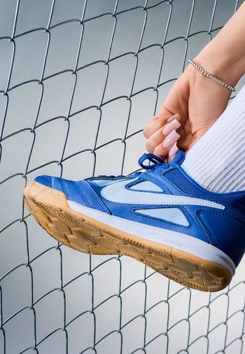 Blaue Sportschuh mit hellblauen Akzenten, strukturiertem Mesh und Gummisohle, vor einem Zaun positioniert.
