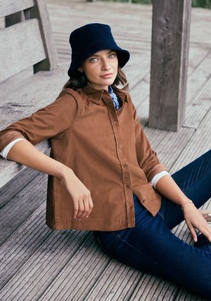 Brown corduroy button-up shirt with a pointed collar, worn over a blue patterned scarf, paired with dark jeans and a navy bucket hat.