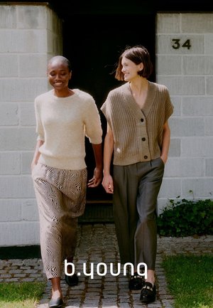 Two women walk together. One wears a light, fuzzy cream sweater and patterned wide-leg pants. The other has a chunky, beige button-up vest and grey trousers.