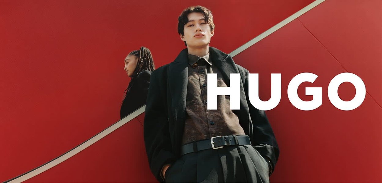 Zwarte jas en overhemd met patroon gecombineerd met high-waisted broek; sterke rode achtergrond met een gebogen witte lijn en de tekst "HUGO".