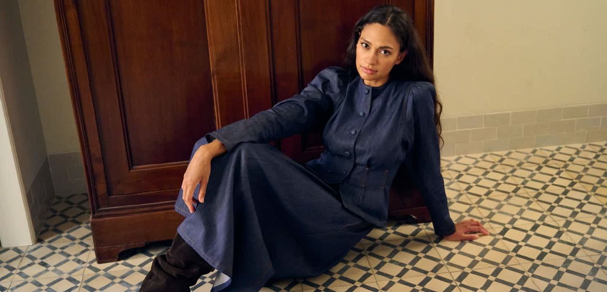 Frau mit langen dunklen Haaren sitzt auf gemustertem Fliesenboden, lehnt an einem Holzschrank und trägt ein dunkelblaues, geknöpftes Kleid sowie schwarze Stiefel.