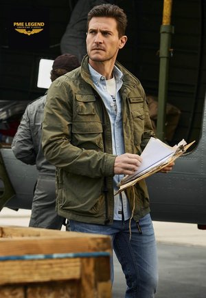 Hombre con chaqueta verde y camisa azul sosteniendo un portapapeles, de pie cerca de un avión con otra persona con monos grises al fondo.