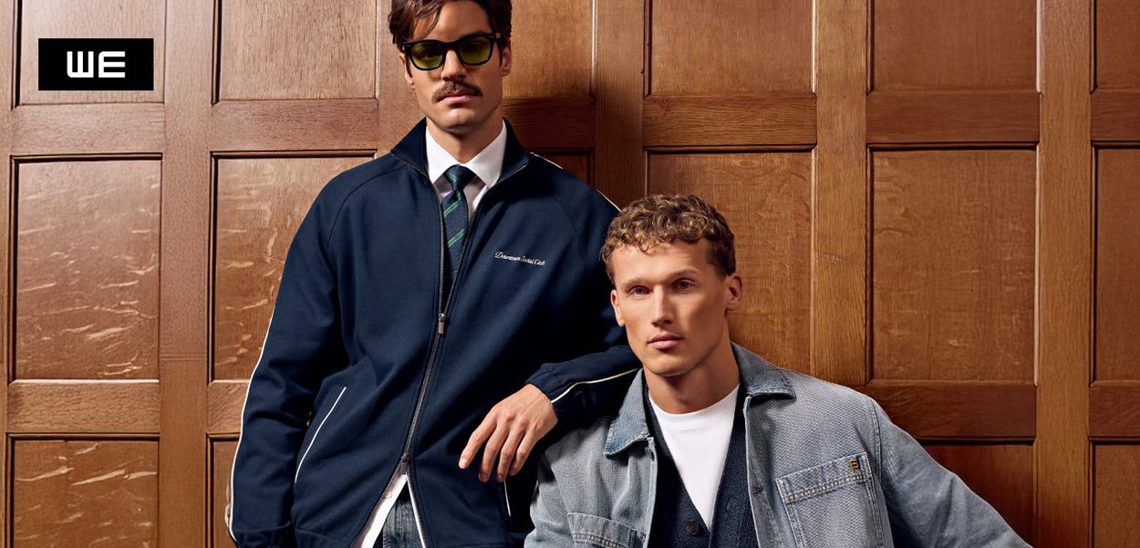 Zwei Männer stehen vor Holztafeln. Einer trägt eine dunkelblaue Jacke, der andere eine Jeansjacke über einem weißen Hemd, beide aus strukturierten Stoffen.