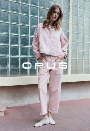 Femme debout contre un mur en blocs de verre portant une veste rose clair et un pantalon taille haute assorti avec des chaussures blanches sans lacets, les mains dans les poches.