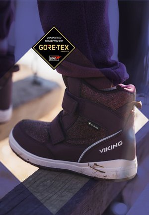 Lila wasserdichte Stiefel mit strukturiertem Obermaterial, Klettverschlüssen und weißen Sohlen. Mit Gore-Tex-Branding an der Seite.