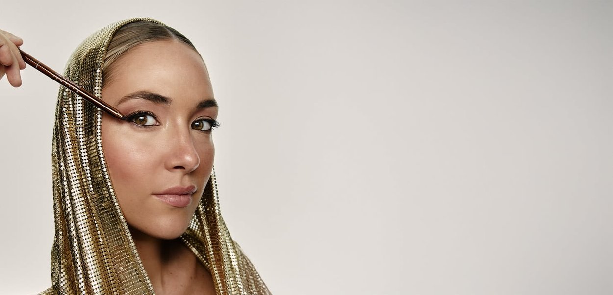 Gold sequined headscarf drapes over hair. The subject applies eyeliner with a brush, showcasing a natural makeup look and smooth skin texture.