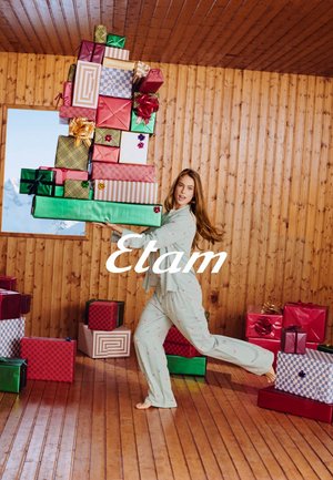 Des cadeaux verts et rouges empilés avec des nœuds colorés ; une personne en pyjama vert clair à motifs les porte, sur fond de bois.