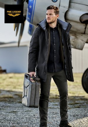 Chaqueta aislante negra con capucha, camisa negra abotonada, pantalones grises y botas negras; sosteniendo un recipiente metálico gris. Avión al fondo.