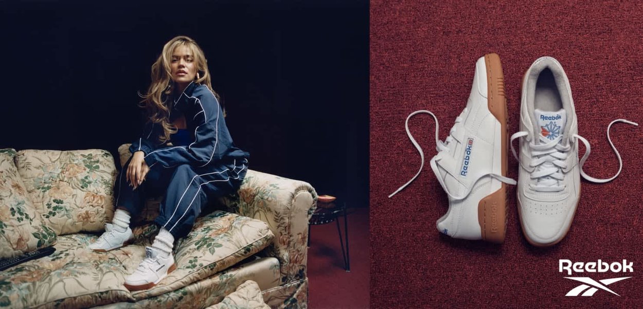 Mujer con chándal azul marino sentada en un sofá floral, lleva zapatillas blancas Reebok junto a un primer plano de zapatillas blancas Reebok sobre una alfombra roja.